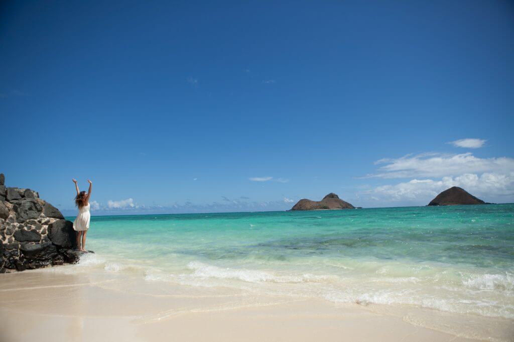 Lanikai Beach is one of the best beaches on Oahu for families. Image of a woman with her arms stretched up standing on the beach overlooking two little islands.