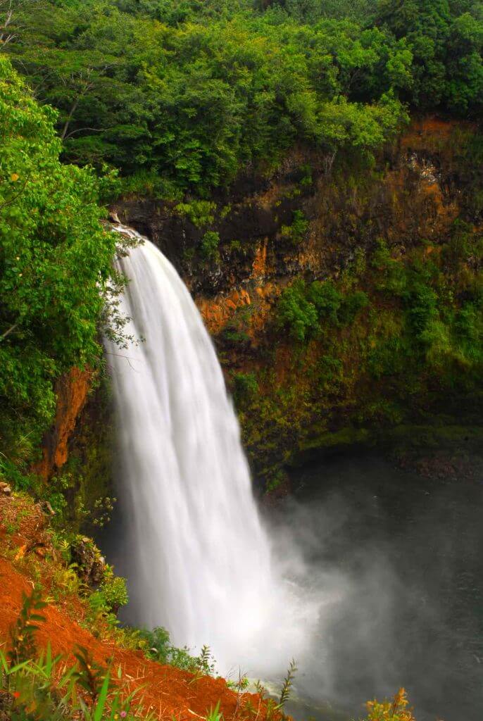 Wailua Falls is one of the most popular waterfalls on Kauai. Image of a gushing waterfall surrounded by greenery and red dirt.