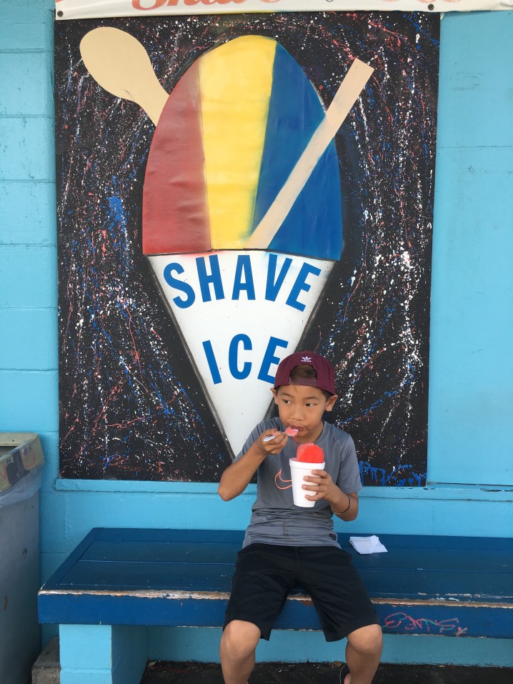 Image of a boy eating shave ice out of a styrofoam cup on a bench in front of a large Shave Ice sign.