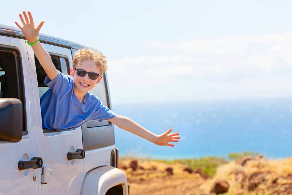 Tips for Renting a Car in Hawaii featured by top Hawaii blog, Hawaii Travel With Kids: happy positive caucasian boy in sunglasses peeking out of the car window with his hands up in the air, tropical family vacation or active road trip concept