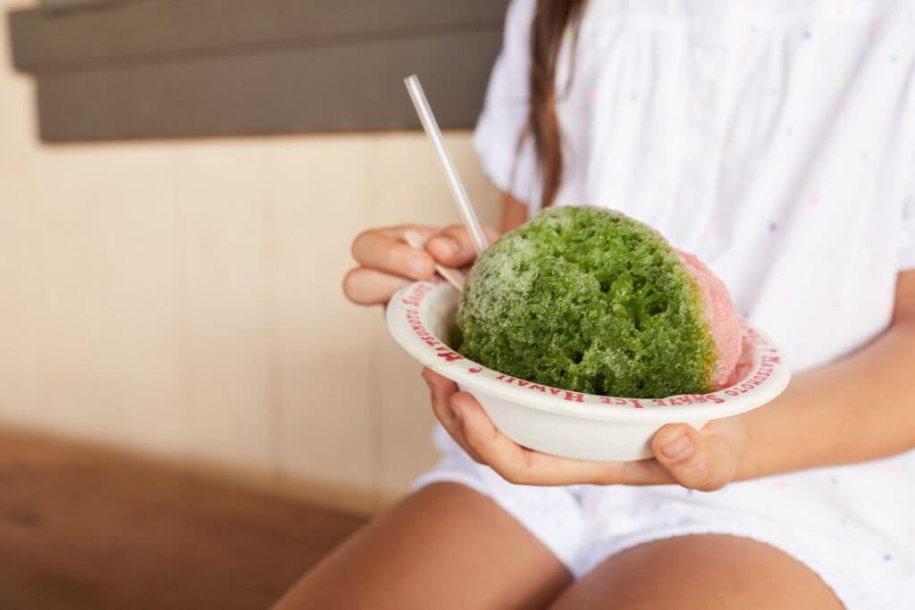 One of the best places to get shave ice on Oahu is Matsumoto's. Image of a woman holding a paper bowl with green and pink shave ice.