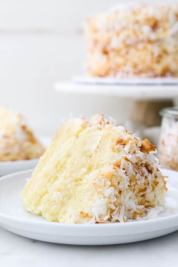 61 Delicious Coconut Dessert Recipes Perfect for Summer featured by top Hawaii blog, Hawaii Travel with Kids: A closeup of a layered coconut cake on a white rimmed plate, the cake stand is blurred out in the background