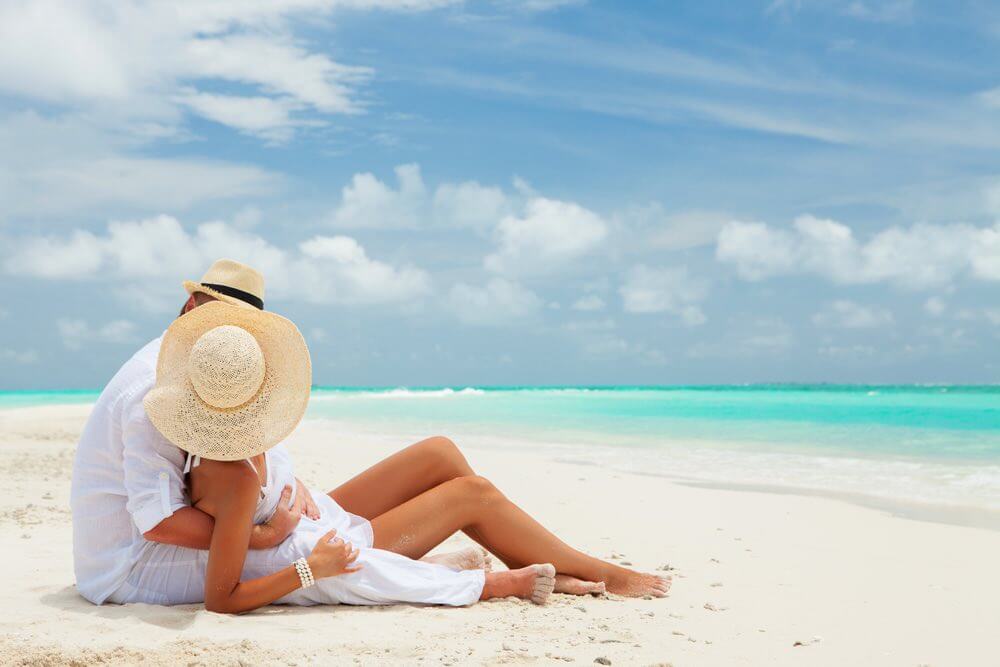 Wondering what's the best island in Hawaii for a honeymoon? It totally depends on what you want to do. Image of a man and woman wearing white looking romantic on a white sandy beach.