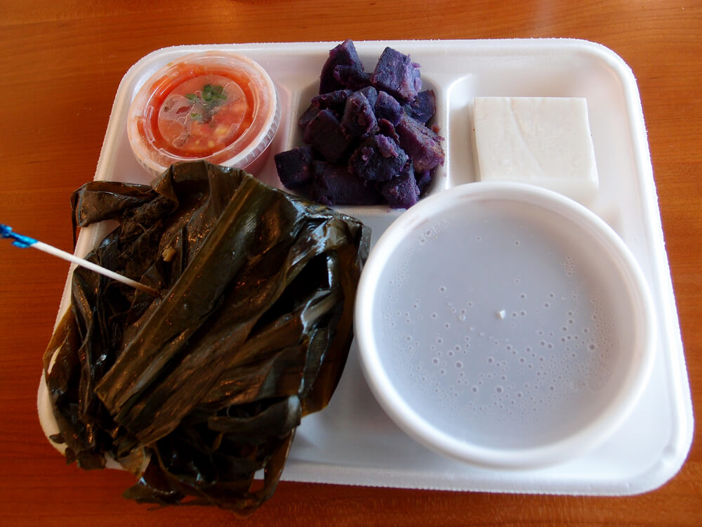 Hawaiian Lau Lau plate Served with Fresh/Day Old Poi in bowl, 'Uala (sweet Potato), Haupia with Lomi Salmon in a plastic container on a Styrofoam plate on a table.