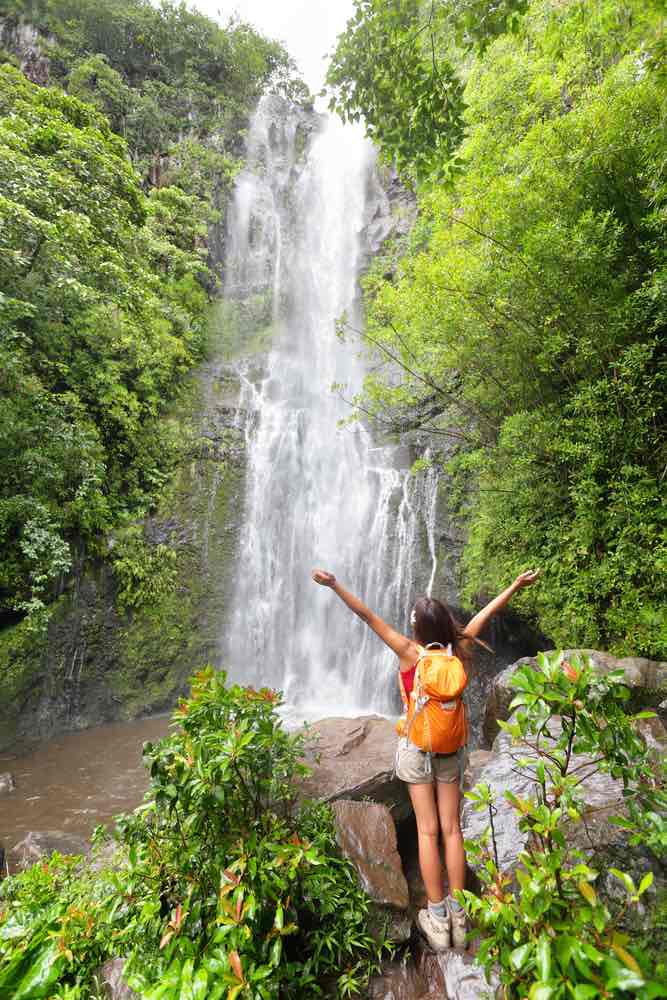 The Road to Hana is a top Maui attraction. Image of a woman wearing an orange backpack standing in front of a waterfall with her arms up.