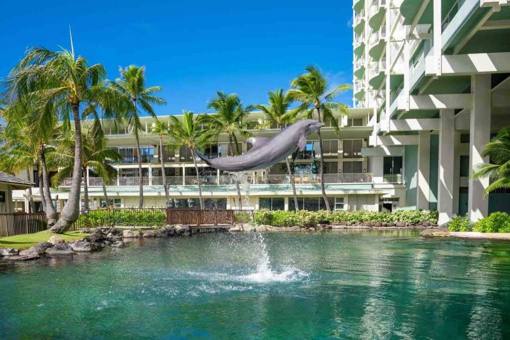 The Kahala Resort and Spa on Oahu is one of the Best Hawaii Hotels for Families. Image of a dolphin jumping in water in front of a hotel.