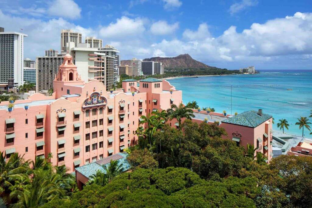 Stay at the Royal Hawaiian Hotel in Waikiki, Oahu with kids. Image of a pink hotel in Waikiki with Diamond Head in the background.
