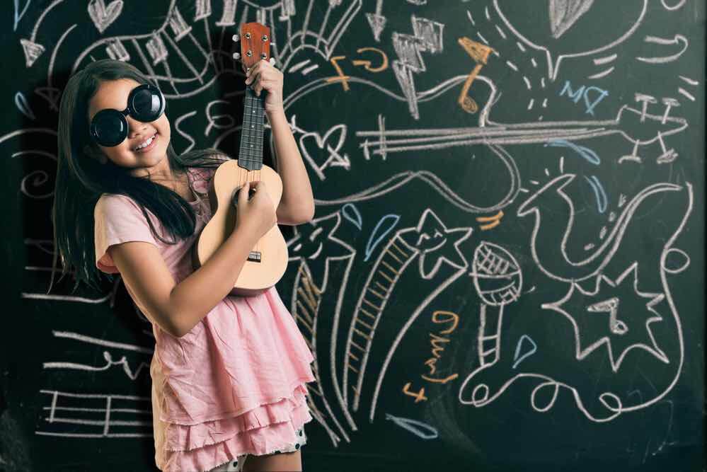 Find out whether you should buy an ukulele for kids in this children's ukulele buying guide. Image of a girl wearing sunglasses and playing the ukulele in front of a chalkboard