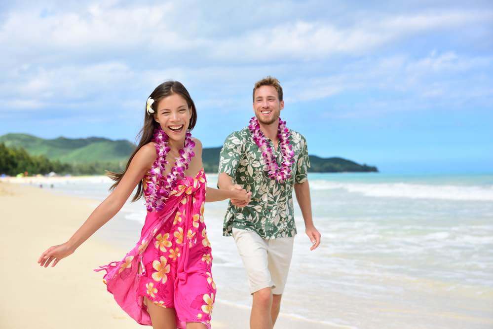 6 Best Kauai Luau Options featured by top Hawaii blogger, Hawaii Travel with Kids: Happy couple having fun running on Hawaii beach vacations in Hawaiian clothing wearing Aloha shirt and pink sarong sun dress and flower leis for traditional wedding or honeymoon concept.