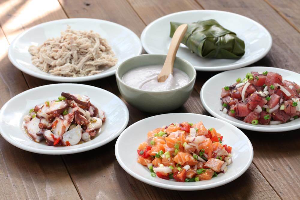 Traditional Hawaiian Food items totally worth trying. Image of white plates of lomi lomi salmon, poke, lau lau, kalua pork, and poi.