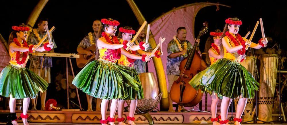 The Ka Wa'a luau is one of the best luaus on Oahu for families. Image of several female hula dancers wearing ti leaf skirts and playing with split bamboo.