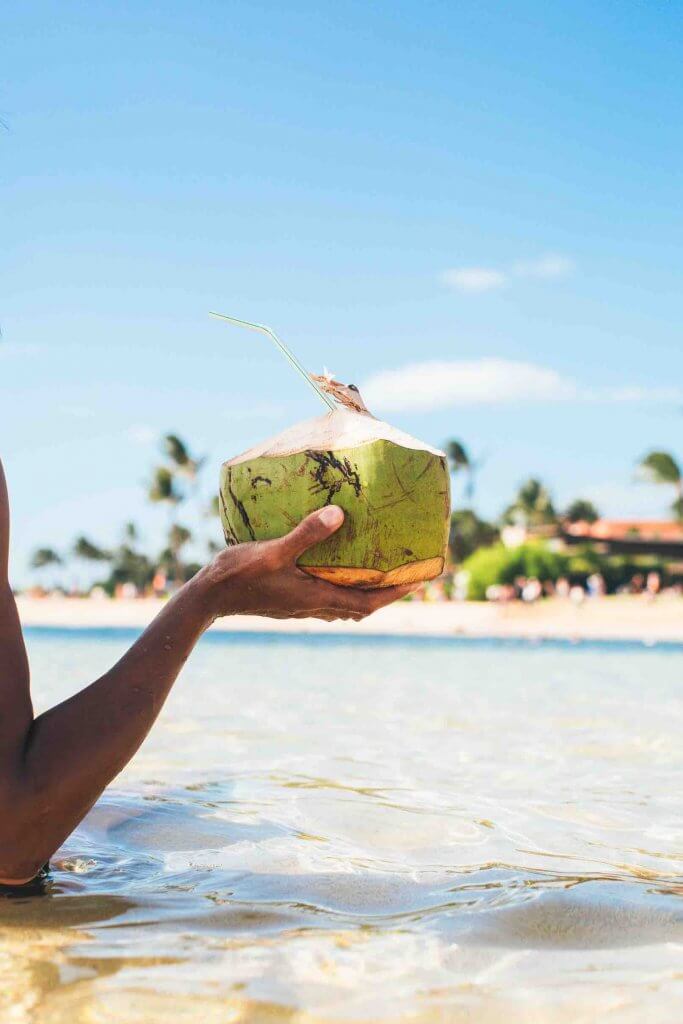 One of the best things to do in Oahu with kids is let them taste fresh coconut water. Image of a woman holding a fresh coconut at the beach.