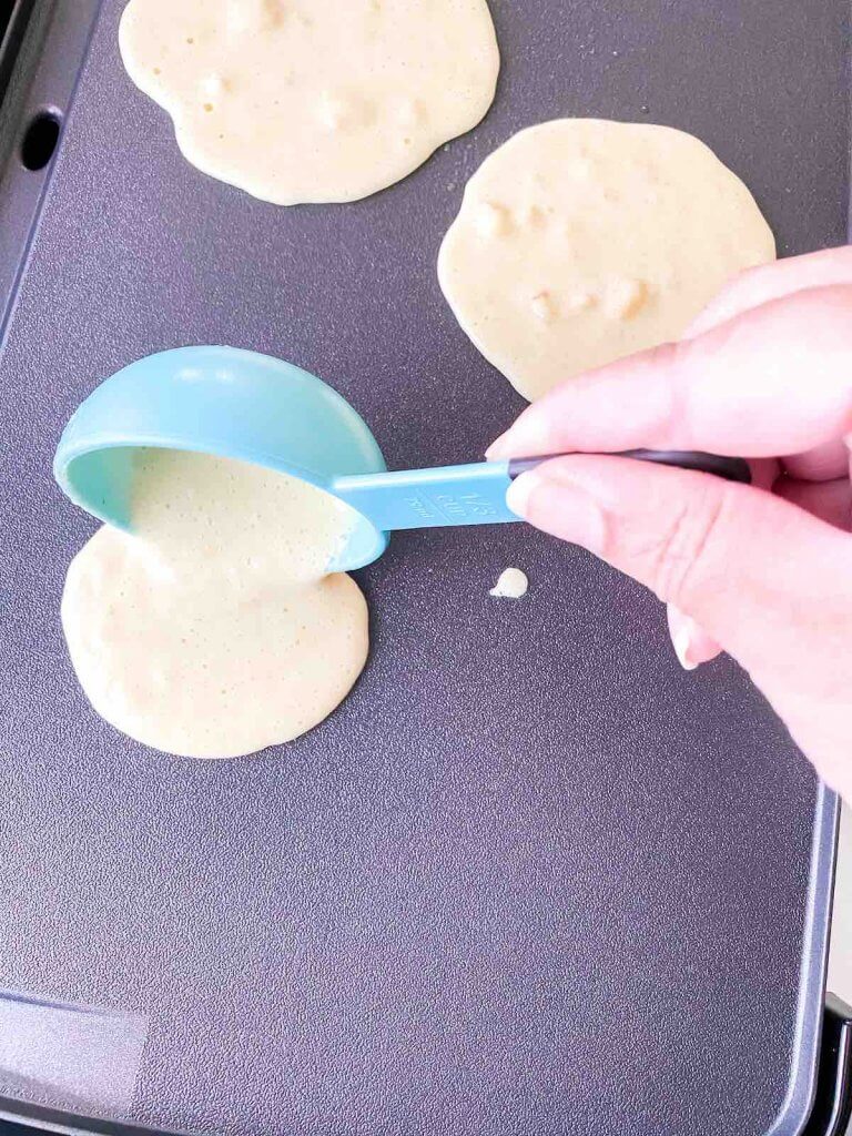 You'll want to use a measuring cup to pour the right amount of gluten free healthy pancakes batter on the griddle. Image of a blue measuring cup pouring Hawaiian pancake batter on the griddle.