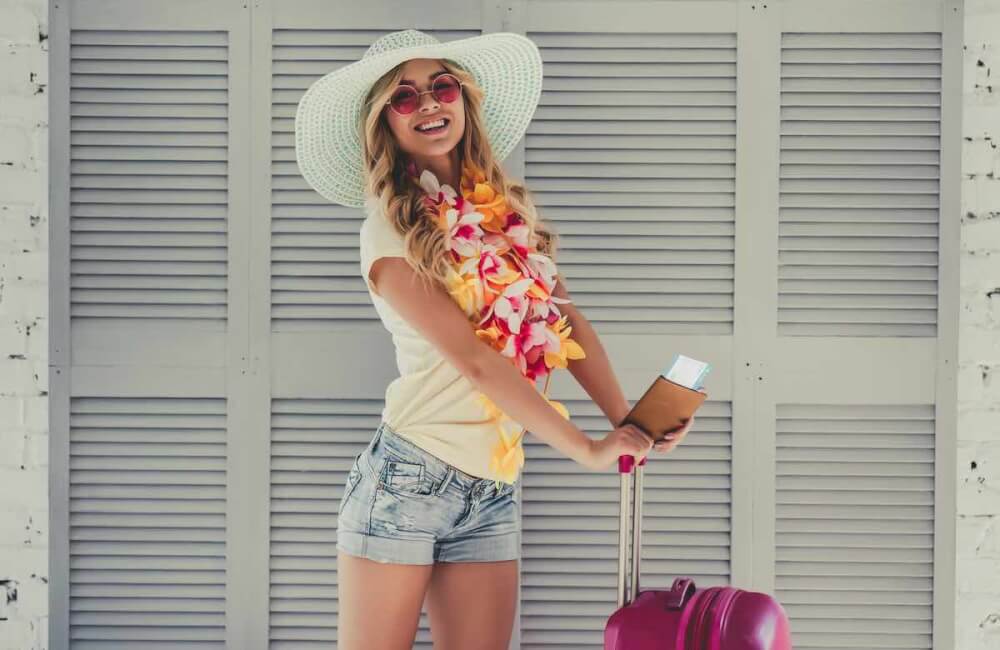 Planning a Hawaii trip and want to know know the best Hawaii travel advice? Find out what you need to know before booking your Hawaii vacation, espeically if it's your first time visiting Hawaii. Image of Attractive young woman is preparing for traveling. Standing on grey background with big suitcase in Hawaii accessories, pink sunglasses and hat with passport and ticket in hand.