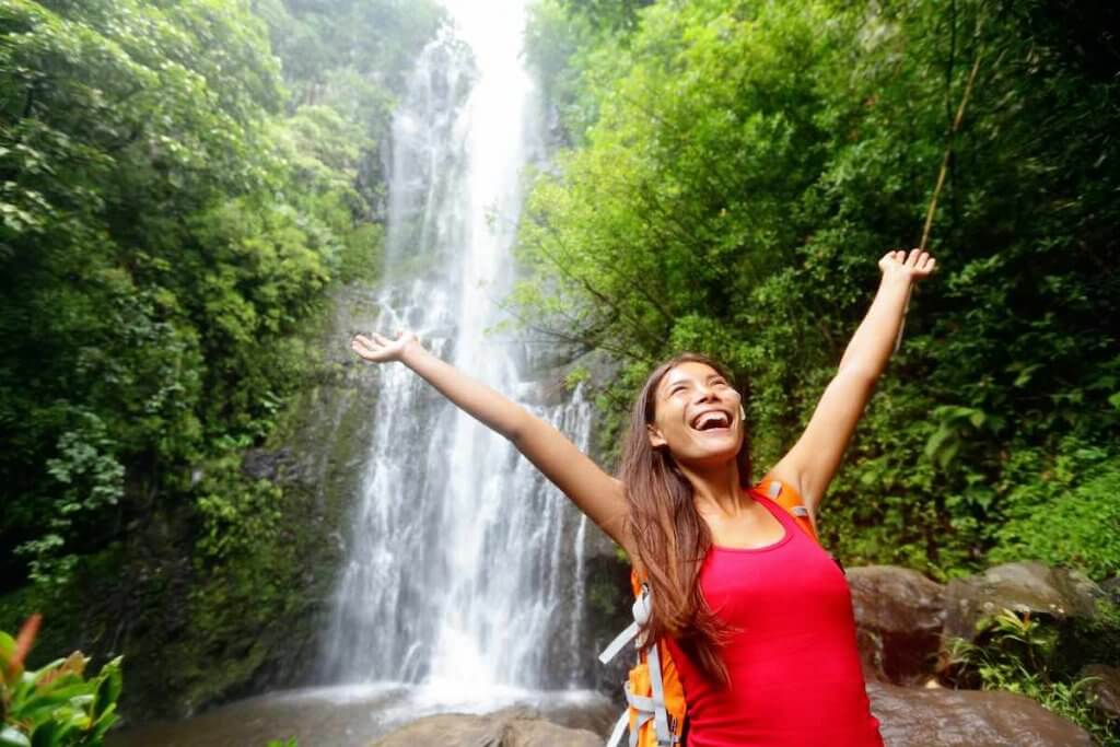 Find out all the best tips for planning a trip to Maui by top Hawaii blog Hawaii Travel with Kids. Image of Hawaii woman tourist excited by waterfall during travel on the famous road to Hana on Maui, Hawaii. Ecotourism concept image with happy backpacking girl. Mixed race Asian / Caucasian backpacker.