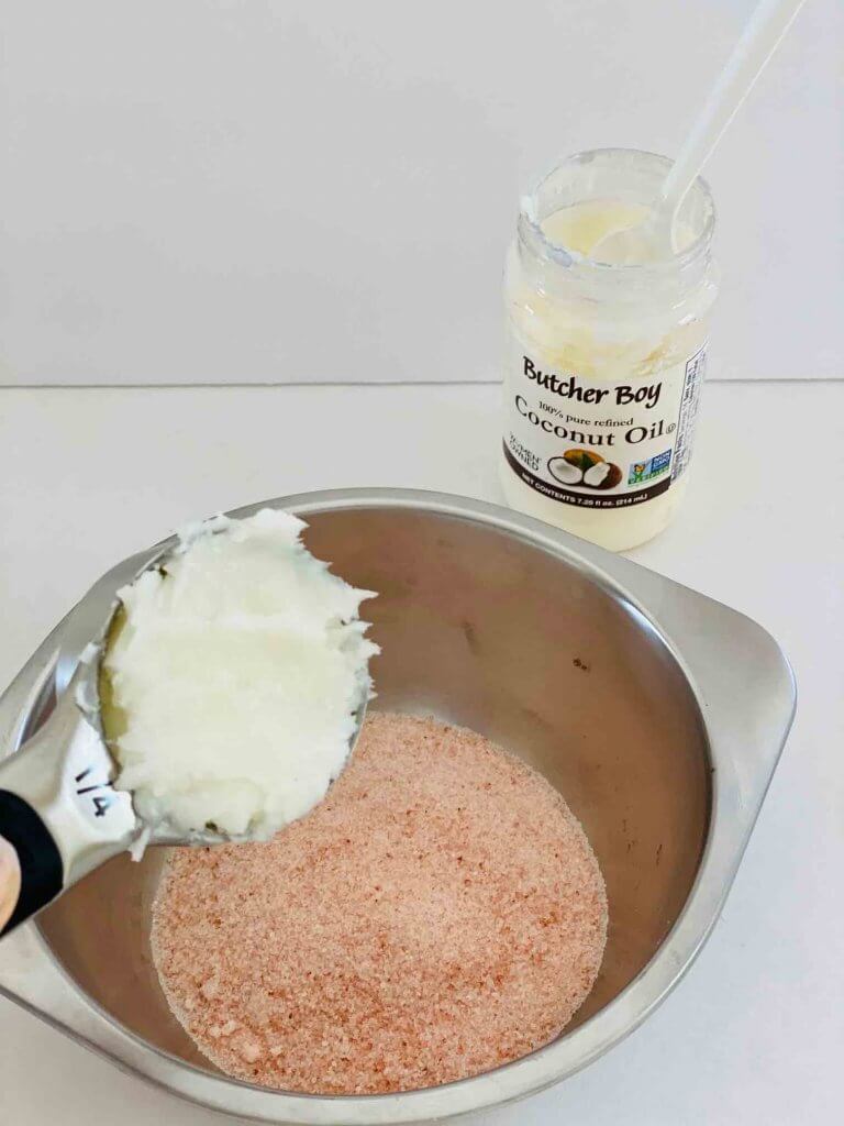 Mix in coconut oil for this homemade coconut oil body scrub. Image of someone measuring coconut oil over a bowl of pink sea salt.