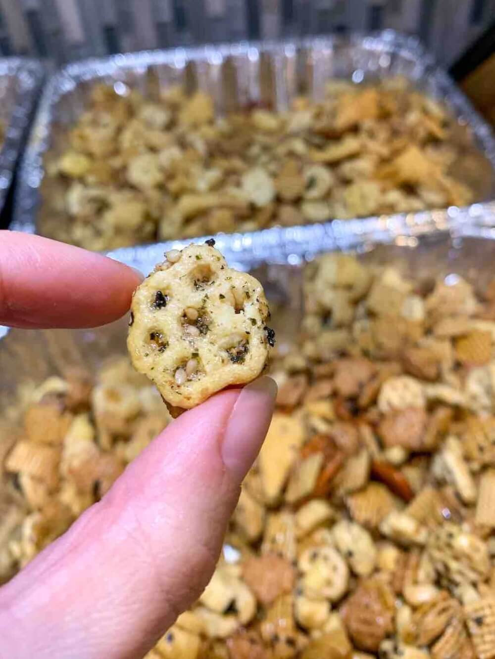 Image of someone holding up a piece of Honeycomb cereal coated in furikake seasoning in front of a pan of Hawaiian Furikake Chex Mix.