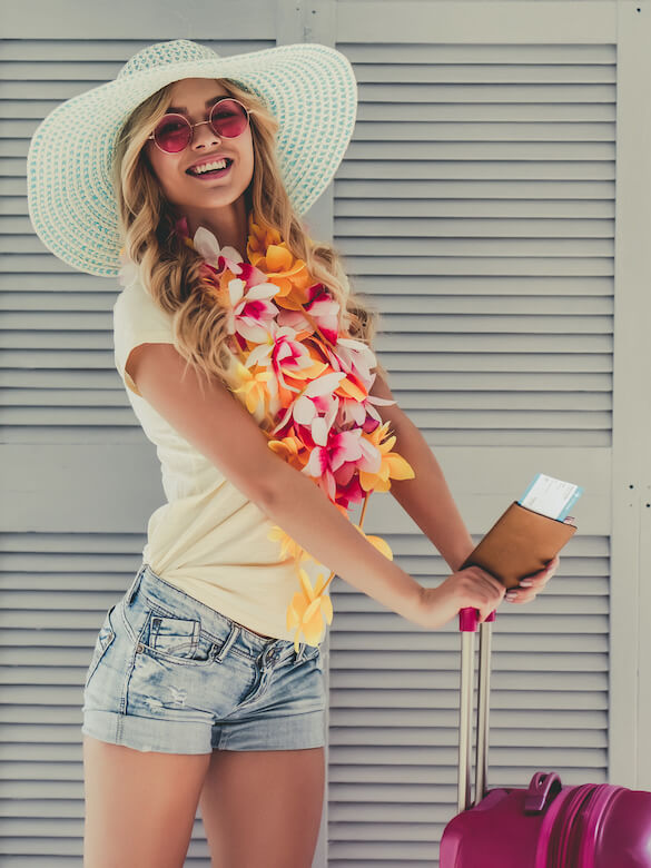 Find out what you might forget on your Hawaii packing list. Image of a woman wearing a large white straw hat, sunglasses, and a lei holding a passport and a pink rolling suitcase, ready for her first trip to Hawaii.