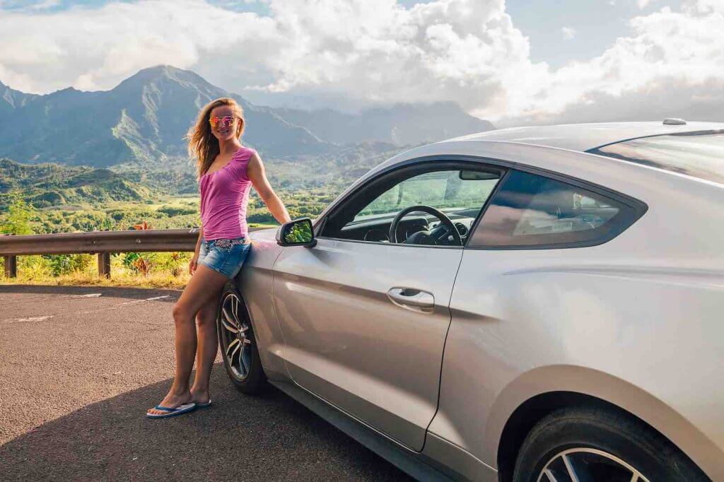 One of my top tips for planning a trip to Kauai is to rent a car. Image of a woman posing by a rental car on Kauai.