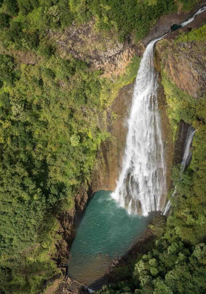 Manawaiopuna Falls is one of the best waterfalls on Kauai. Image of Manawaiopuna Falls on Kauai going into a blue pool surrounded by lush greenery.