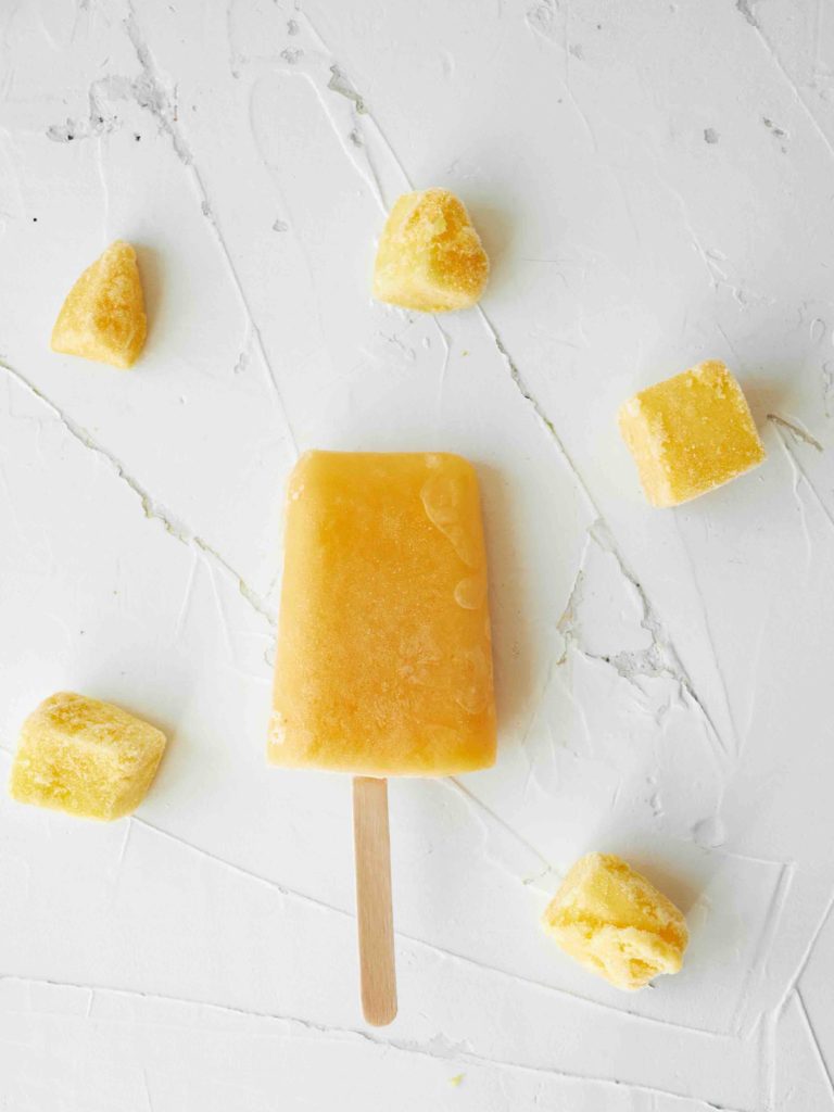 Learn how to make mango popsicles by top Hawaii blog Hawaii Travel with Kids. Image of a homemade mango popsicle surrounded by frozed mango chunks on a white background.