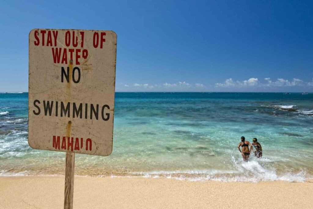 Image of a sign that reads "stay out of water, no swimming, Mahalo" with two people in the water in Hawaii.