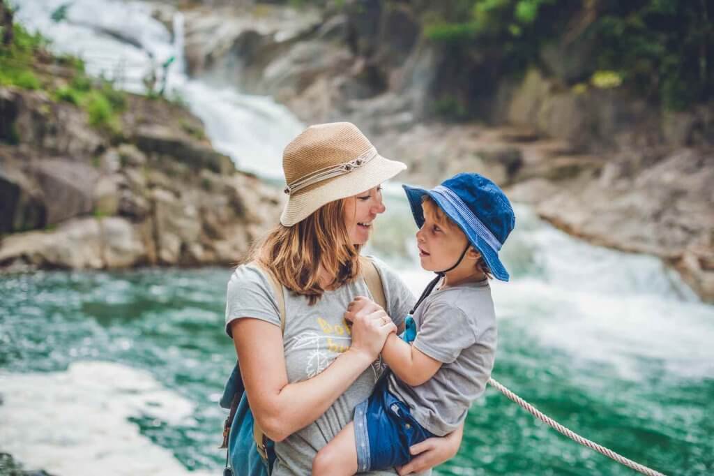 Find out the best things to do in Hawaii with toddlers by top Hawaii blog Hawaii Travel with Kids. Image of Mother and son relaxing under a waterfall. Vacation concept.