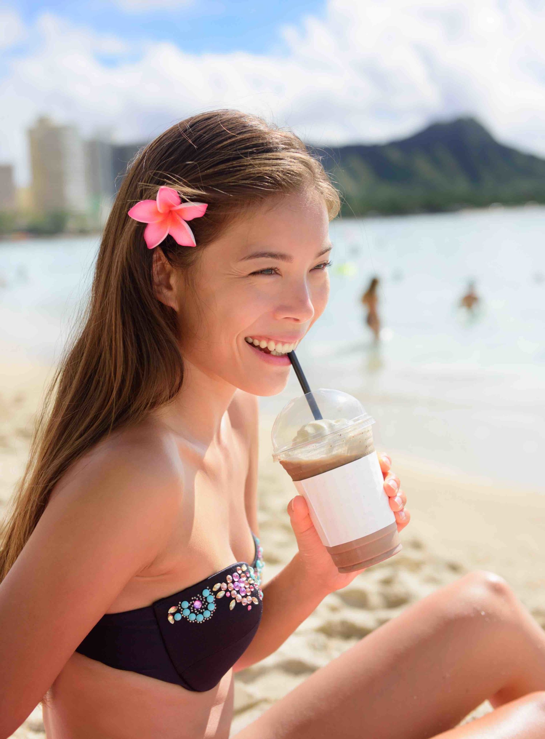 Find out all the best things to do in Waikiki Oahu by top Hawaii blog Hawaii Travel with Kids. Image of a woman drinking an iced coffee while sitting on Waikiki Beach with Diamond Head in the background.