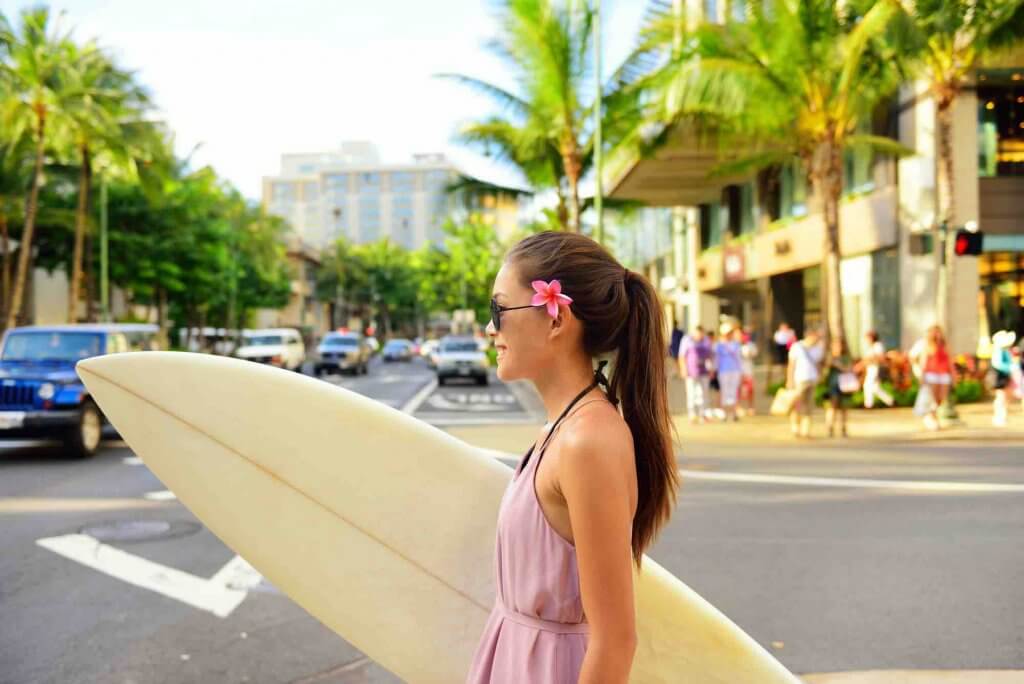 Find out the best Oahu planning tips from top Hawaii blog Hawaii Travel with Kids.Image of Surfer woman walking in city with surfboard to go surfing. Urban Hawaiian surf concept. Asian girl holding surf board crossing street to go to the beach. Waikiki, Honolulu city, Oahu, Hawaii, USA.