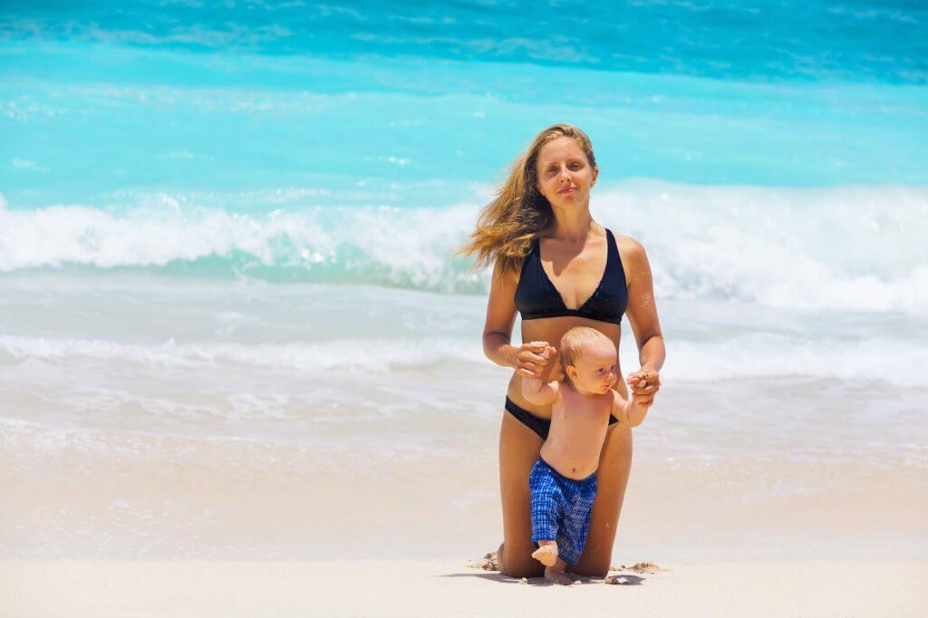 Read this baby-friendly Hawaii guide by top Hawaii blog Hawaii Travel with Kids. Image of On sunny white sand beach happy young mother hold on hands little baby son, have fun before swimming in sea. Active children, travel lifestyle, summer vacation with kids on tropical family resort.