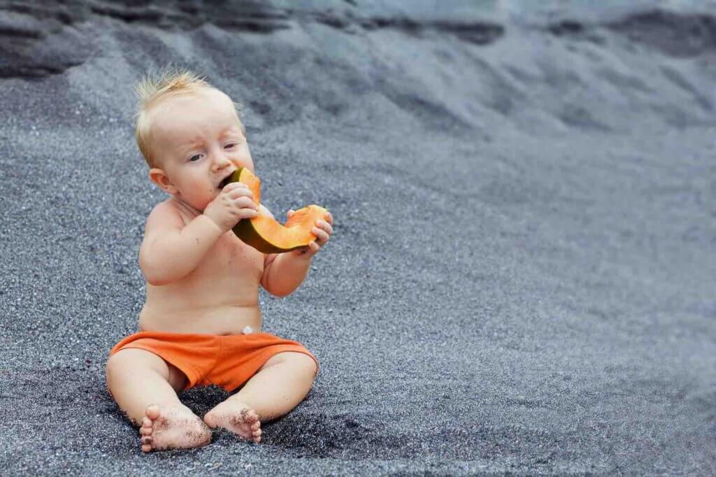 There are tons of things to do with a baby in Hawaii. Image of a baby boy with smiling face eating ripe fruit - orange papaya slice. Healthful food, natural breakfast on sea black beach. Healthy lifestyle on summer family holiday with kids.