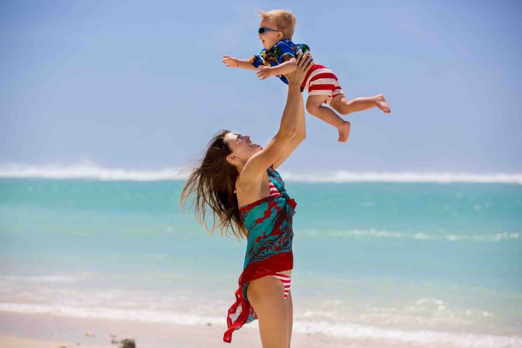 Find out how to travel to Hawaii with a toddler. Image of a mom and child, dressed in Hawaiian shirts, playing together on the beach in Hawaii.