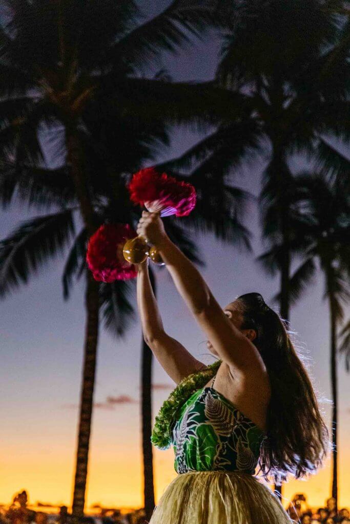 One of the best things to do on Oahu with kids is check out a free hula show. Image of a hula dancer using feather gourd rattles as she dances at sunset.