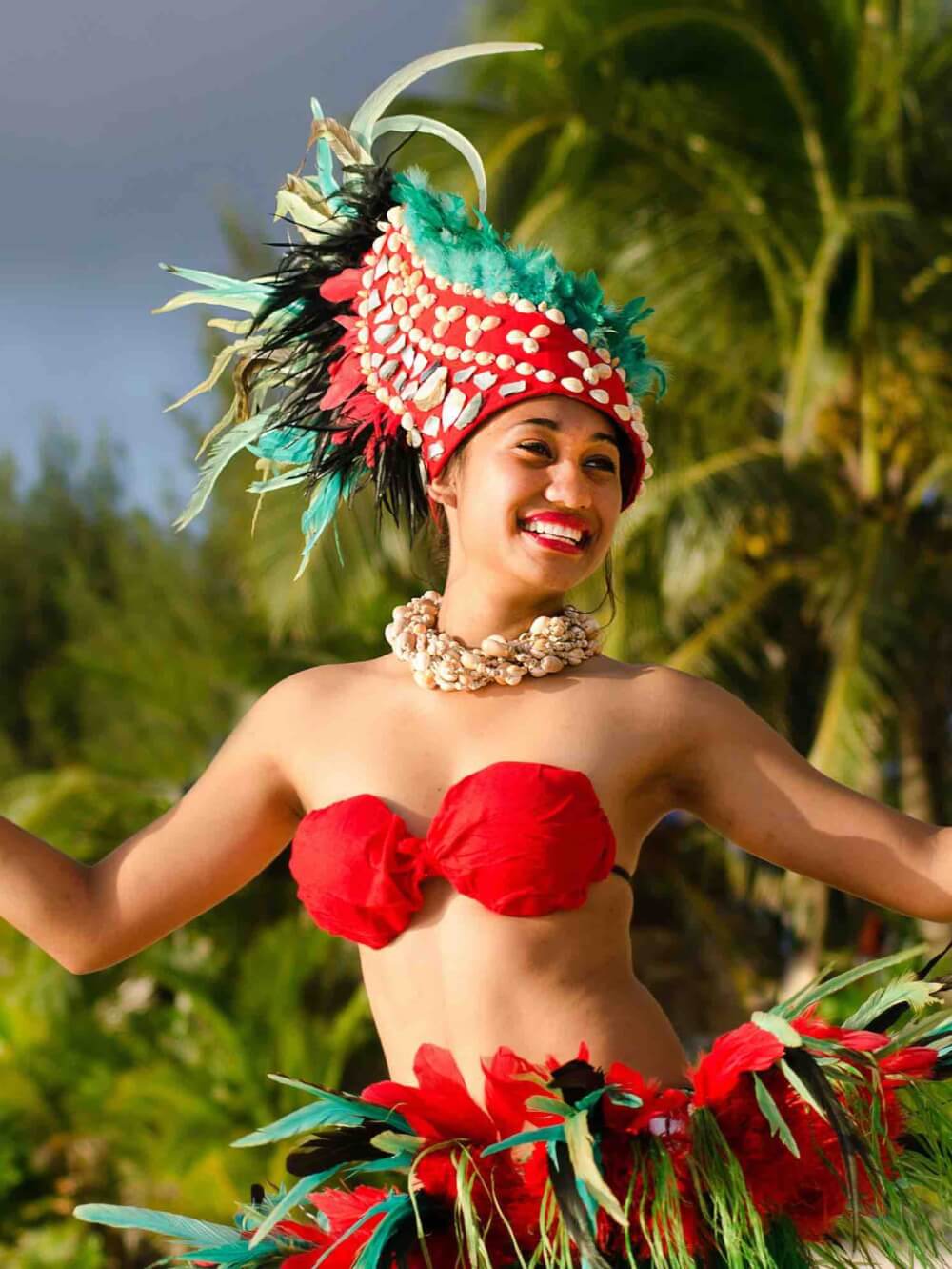 Get all your questions about luaus in Hawaii answered by top Hawaii blog Hawaii Travel with Kids. Image of a Polynesian Pacific Island Tahitian female dancer in colorful costume dancing on tropical beach with palm trees in the background.