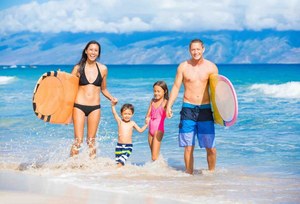 Find out whether Maui vs Oahu is the best Hawaiian island for kids. Image of a family with surfboards on the beach in Hawaii.