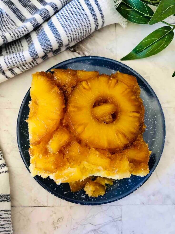 Learn how to make pineapple upside down rum cake by top Hawaii blog Hawaii Travel with Kids. Image of a square slice of pineapple upside down cake on a plate with a blue and white striped towel and some leaves in the background.