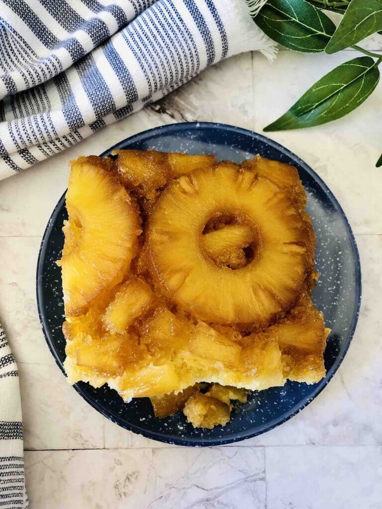 Learn how to make pineapple upside down rum cake by top Hawaii blog Hawaii Travel with Kids. Image of a square slice of pineapple upside down cake on a plate with a blue and white striped towel and some leaves in the background.