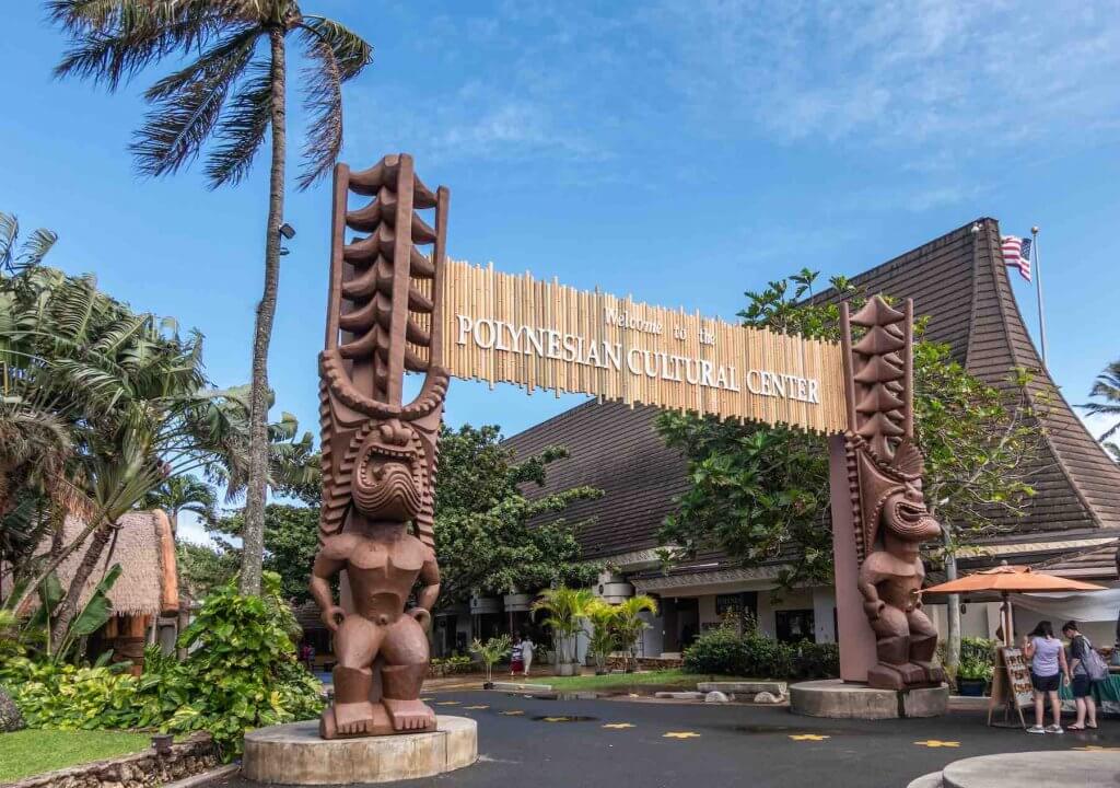 The Polynesian Cultural Center is one of the top Oahu tourist attractions. Image of the Polynesian Cultural Center gate into the park with brown giant tiki statues. People and green vegetation under blue sky.