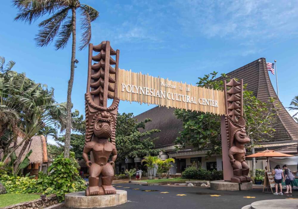 Laie, Oahu, Hawaii, USA. - January 09, 2020: Polynesian Cultural Center. Monumental gate into the park with brown giant aboriginal statues. People and green vegetation under blue sky.