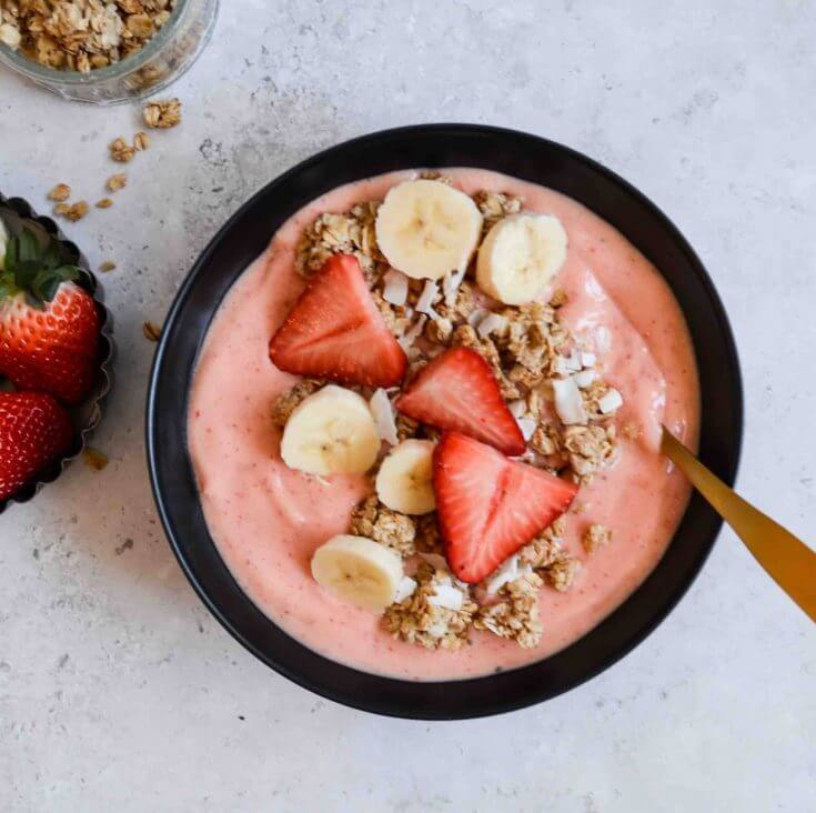 Learn how to make this Strawberry Mango Smoothie Bowl by top Hawaii blog Hawaii Travel with Kids. Image of a pink smoothie bowl topped with fresh strawberris, sliced banana, and granola.