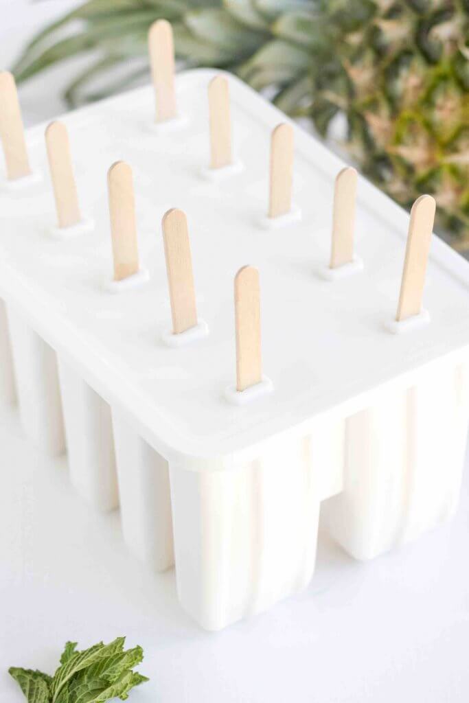 Use these popsicle molds to make these mango popsicles. Image of a white popsicle mold tray next to a fresh pineapple