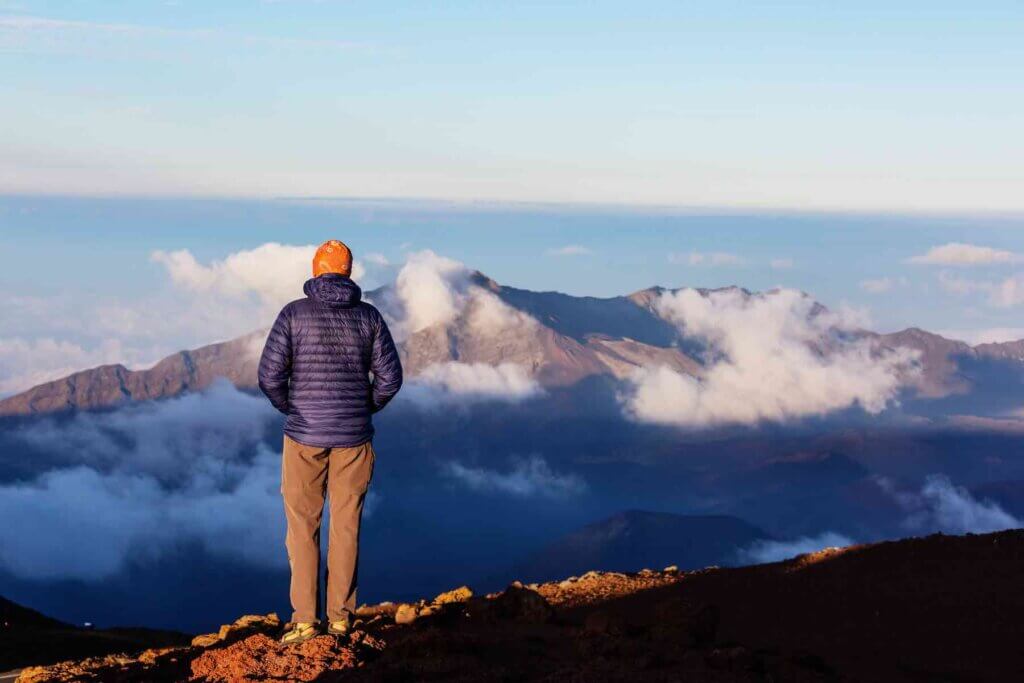 One of the best sunrises on Maui is at Haleakala National Park. Image of a man wearing a hat and coat standing at Haleakala Crater for sunrise.