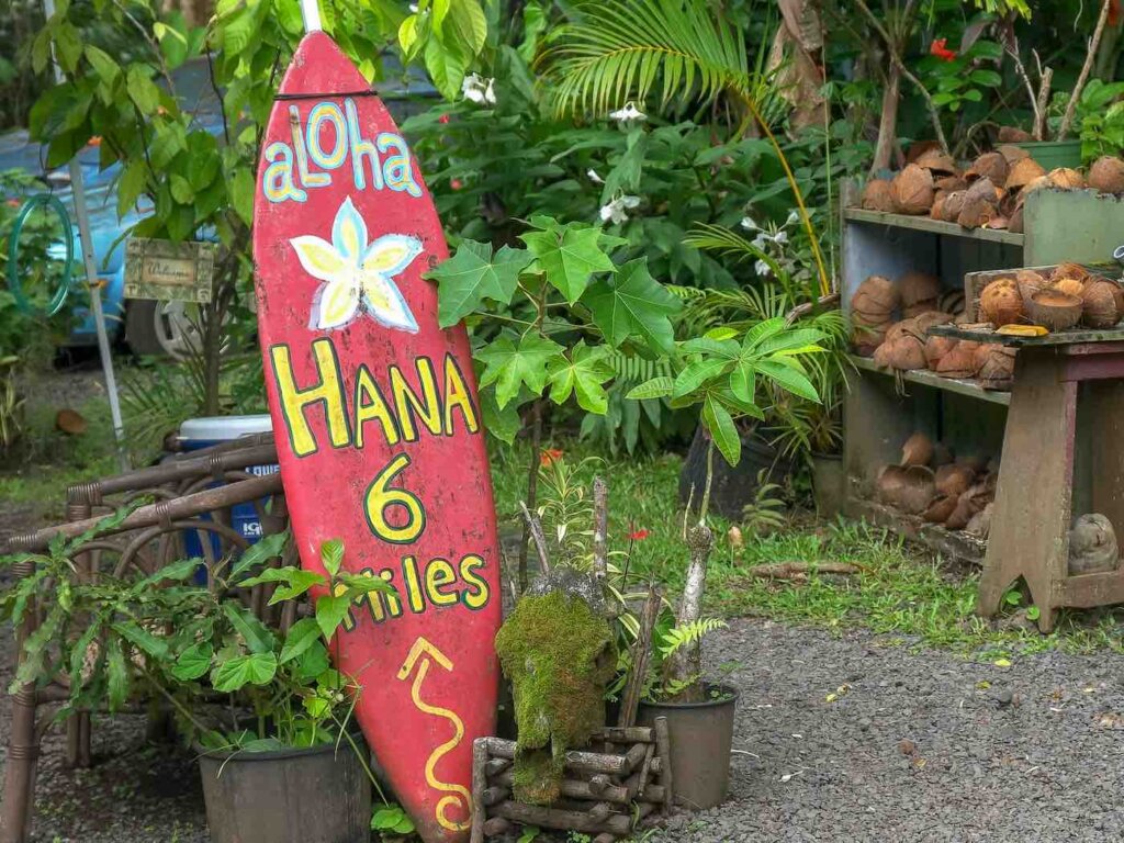 Another fun thing to do in Maui with kids is check out the Road to Hana. Image of a red surfbarod sign that says Hana 6 miles and some fruit in the background.