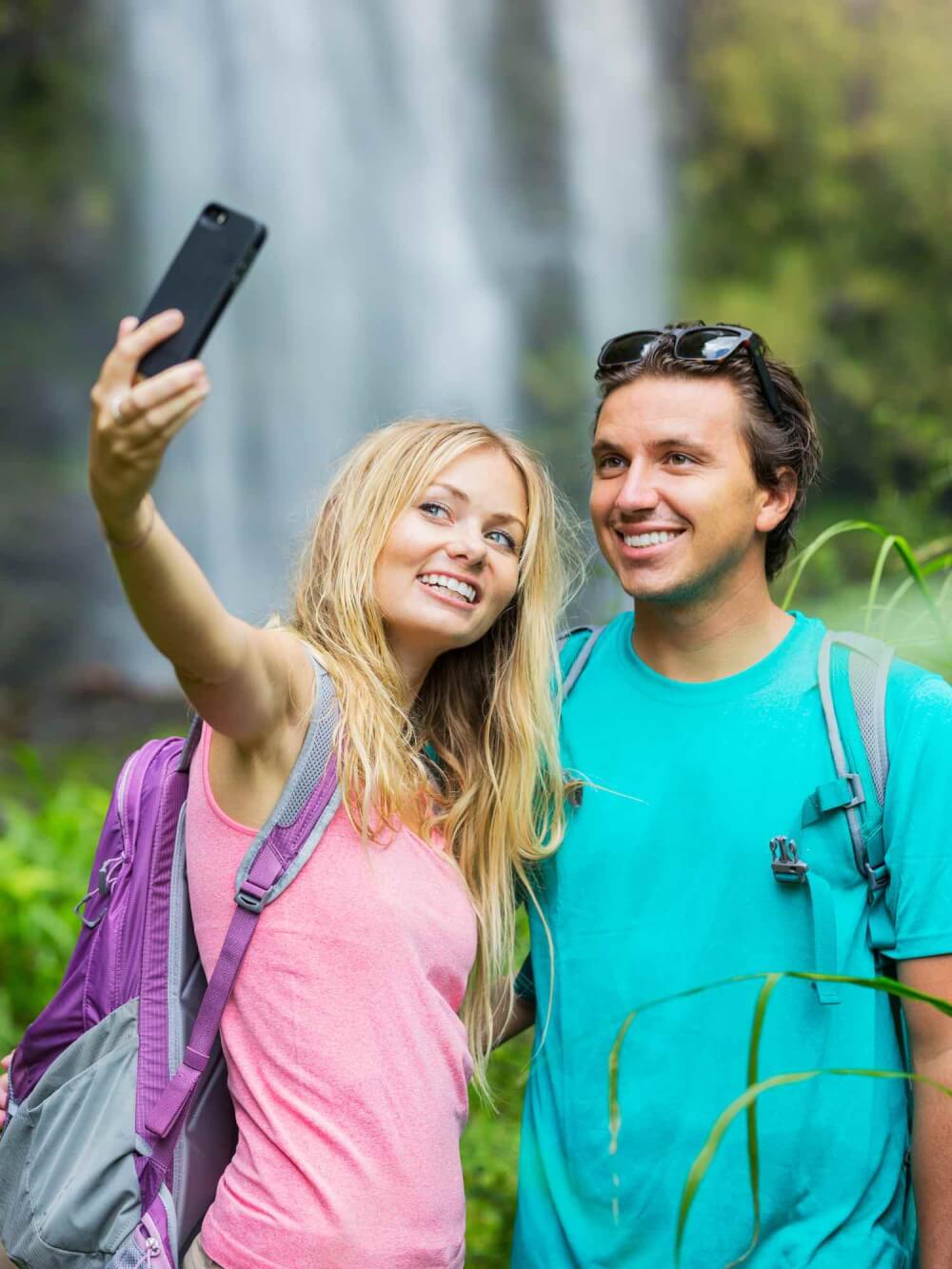 Find out the biggest Hawaii mistakes tourists make in this list by top Hawaii blog Hawaii Travel with Kids. Image of Couple having fun together outdoors. Taking self portrait with camera phone after hiking to incredible waterfall in Hawaii.