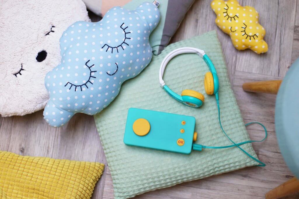 Lunii is the best airplane toy for toddlers. Image of a blue and yellow rectangular box and headphones on a bunch of pillows.