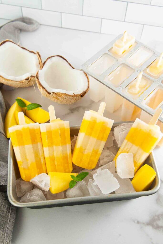 Don't this mango coconut popsicles look tasty? Image of 4 coconut mango popiscles sitting in a tray of ice next to a fresh coconut.