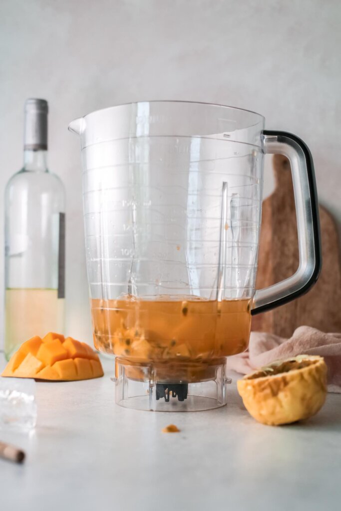 To make a wine slushy, pour all the ingredients into the blender. Image of a blender with fresh mango, passion fruit, wine, and honey inside.