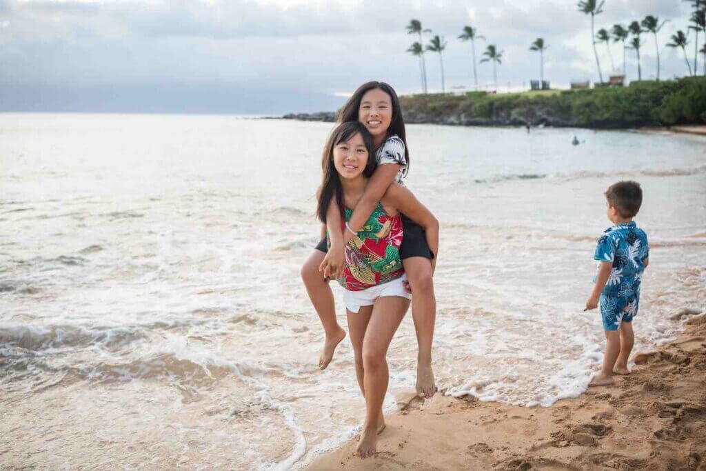 There are tons of tips for geteting awesome Maui family photos with kids. Image of two girls doing a piggy back ride with a little boy in the background