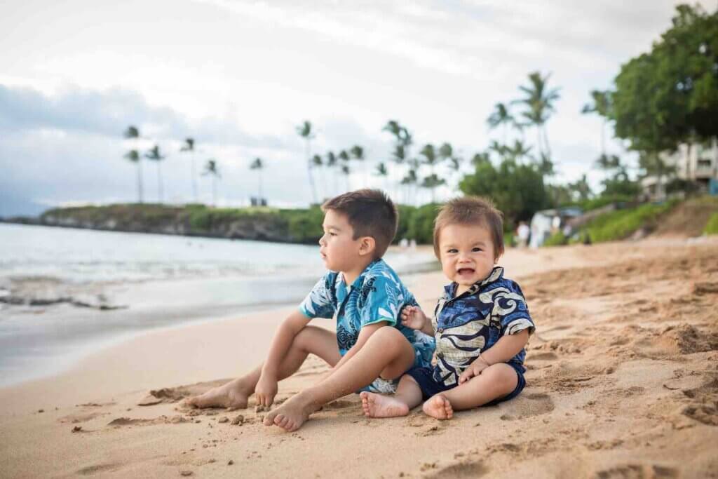 Get cute Maui family photos on your next trip to Hawaii with kids! Image of two young boys wearing Aloha shirts sitting barefoot on a Maui beach.