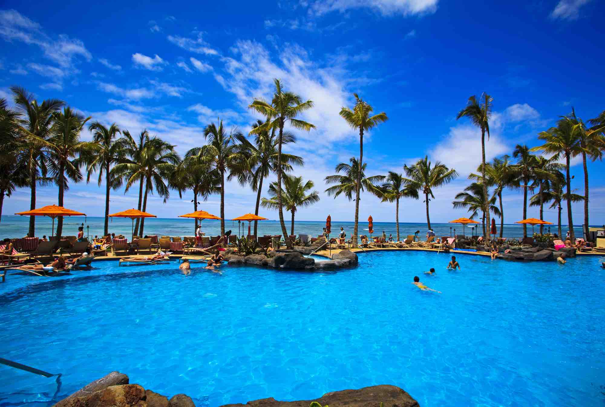 Find out the best resorts in Oahu for families recommended by top Hawaii blog Hawaii Travel with Kids. Image of a resort pool with palm trees and the ocean in the background.